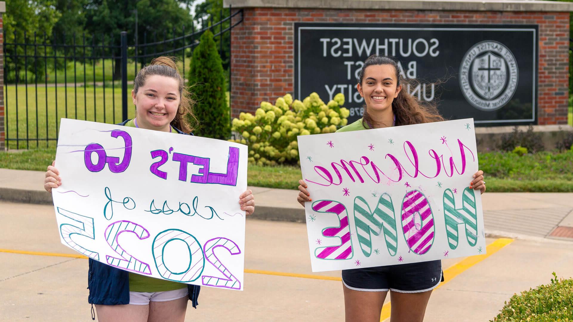 students holding poster board signs that say welcome home while standing in front of S-B-U front entrance